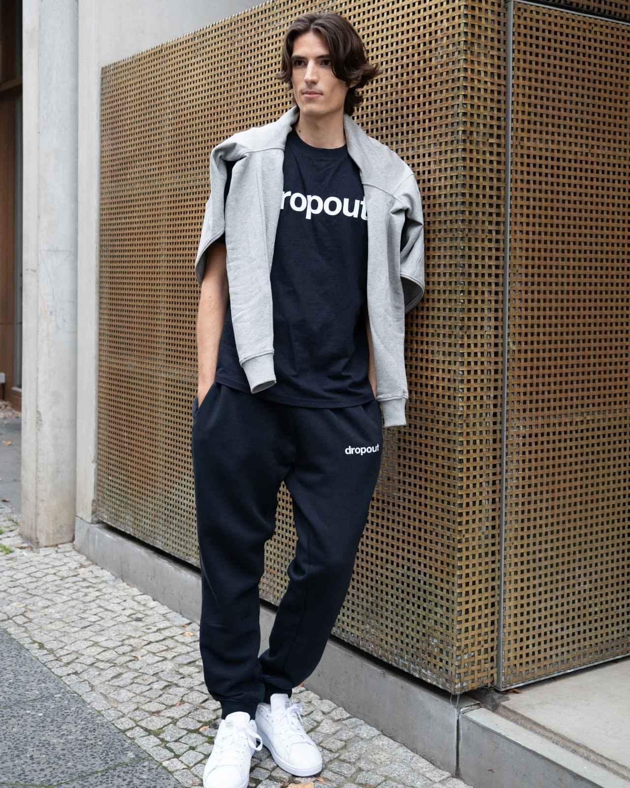 Person wearing a navy 'dropout' t-shirt and pants with a gray jacket against a textured wall.