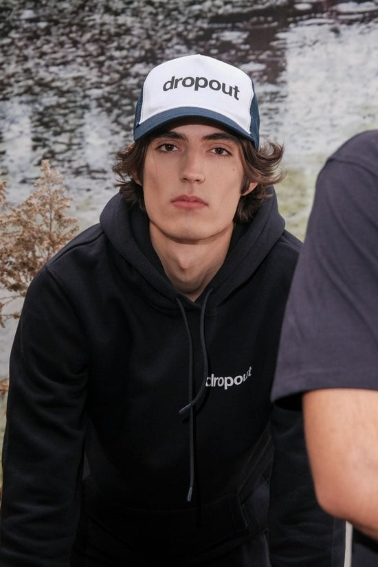 Person wearing a black hoodie and White/Navy cap with 'dropout' branding, sitting outdoors.