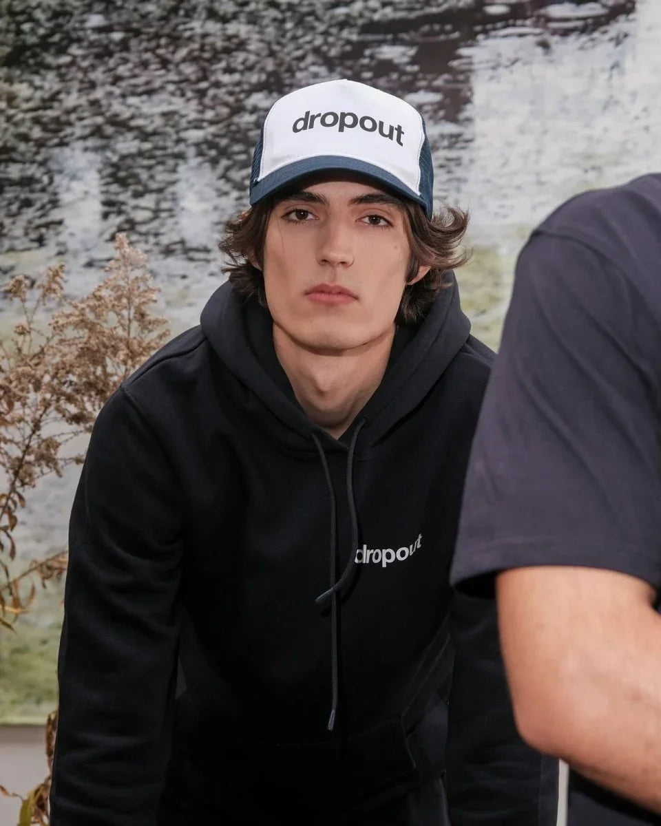 Person wearing a black hoodie and White/Navy cap with 'dropout' branding, sitting outdoors.
